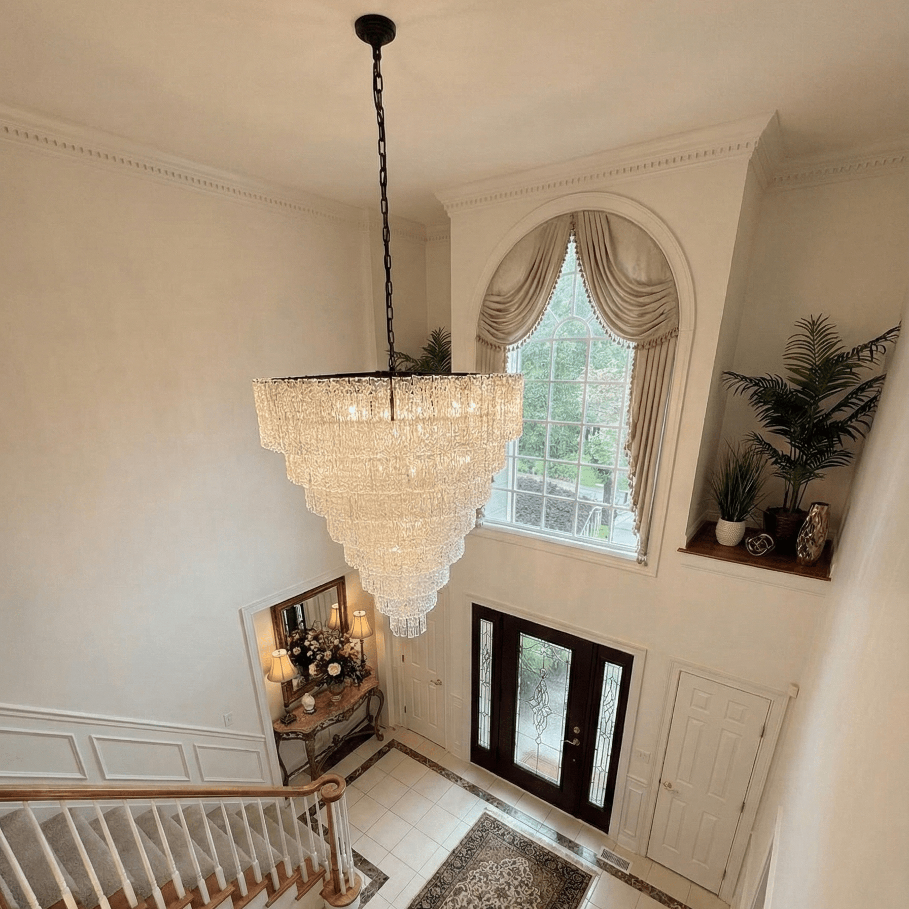 Large tiered crystal chandelier hanging in a grand two-story foyer above a staircase, illuminating a luxury entryway with classic architectural details