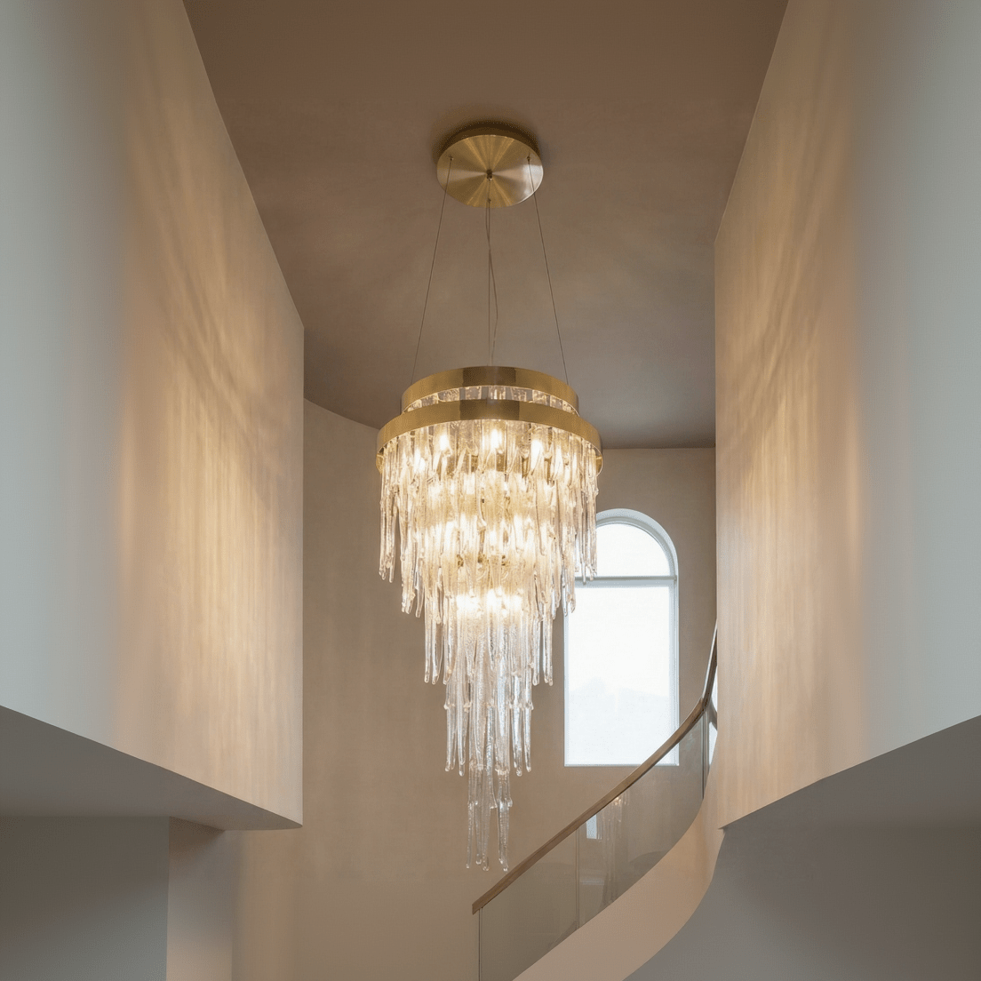 Tall brass tiered crystal chandelier installed in a double-height foyer above a curved staircase, featuring cascading glass icicle drops and warm ambient lighting.